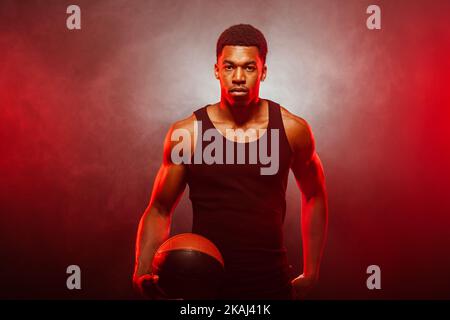 Côté joueur de basket-ball éclairé de couleur rouge tenant une balle sur fond de fumée. Sérieux homme afro-américain concentré. Banque D'Images