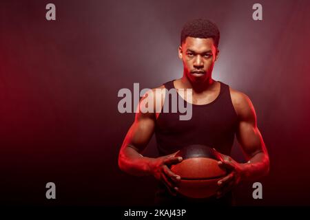 Côté joueur de basket-ball éclairé de couleur rouge tenant une balle sur fond flou. Sérieux homme afro-américain concentré. Banque D'Images