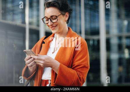 Une heureuse femme d'affaires souriant sur son smartphone tout en se rendant au travail en ville. Femme d'affaires mûre lisant un message texte sur son chemin à l'extérieur Banque D'Images