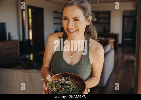 La femme végétalienne a le sourire et regarde loin tout en tenant un bol de bouddha. Bonne femme âgée qui apprécie une salade de légumes après avoir fait de l'activité à la maison. Maturité Banque D'Images