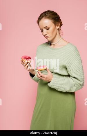 Femme enceinte pensive tenant des beignets doux isolés sur rose, image de stock Banque D'Images