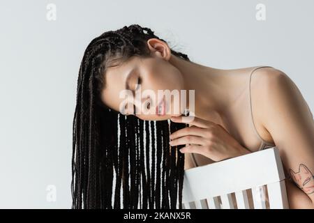 Femme tatouée avec des tresses touchant le menton près de la chaise isolée sur image grise Banque D'Images