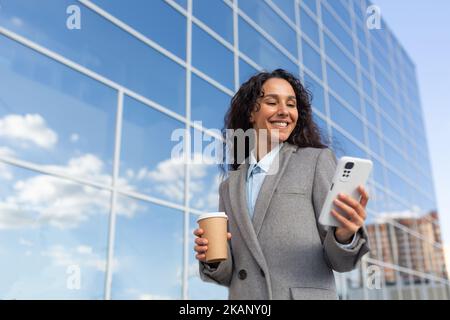 Une femme d'affaires prospère avec un smartphone se rend au travail le matin au bureau, une femme hispanique aux cheveux bouclés souriant et parcourant les actualités en ligne à l'extérieur avec une boisson chaude pour tasse, dans un manteau chaud. Banque D'Images