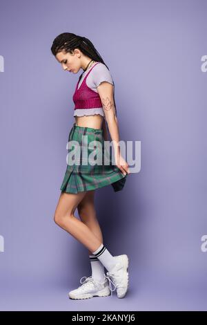 vue latérale de la femme tatouée en jupe à carreaux et baskets blanches debout sur fond violet, image de stock Banque D'Images