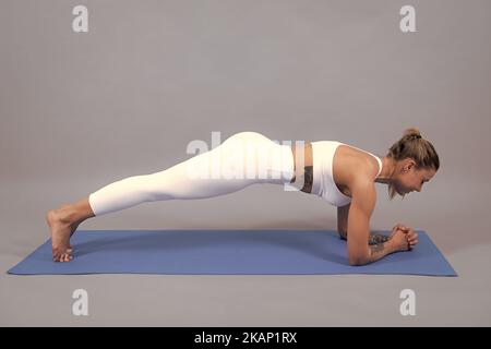 Jeune femme athlétique faisant du planque. Femme de yoga prend des cours de yoga. Une jeune fille sportive paisible dans des vêtements de sport qui s'exercent sur un tapis. Gris isolé Banque D'Images