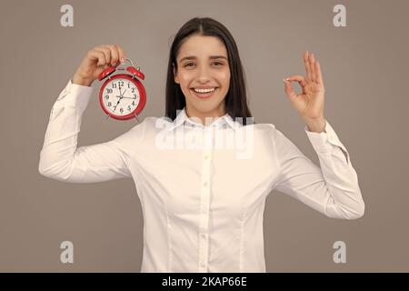 Fille tenant un réveil montre. Portrait d'une jeune femme drôle et belle avec un réveil debout sur fond gris. Banque D'Images