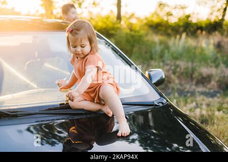 Enfant petit enfant bébé fille assis seul sans surveillance sur le capot de la voiture. Un enfant souriant et heureux. Bonne famille, enfance. Vacances d'été Banque D'Images