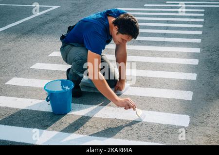 Le préposé à l'entretien routier peint les passages pour piétons rayés sur de l'asphalte à l'aide d'une brosse à peinture. Marquages pour piétons sur la chaussée. Transition sécuritaire pour les personnes. Flux de travail réel... Banque D'Images