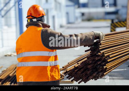 Le Slinger dans le casque de construction et le gilet orange déchargent la cargaison sur le chantier de construction. Vue de l'arrière. Un vrai travailleur. Flux de travail authentique... Banque D'Images