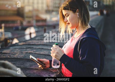 Une jeune femme adulte, plutôt ajustée aux courbes, qui tient une bouteille d'eau, prend une pause après son entraînement et vérifie ses messages sur son téléphone portable en portant des écouteurs - Banque D'Images