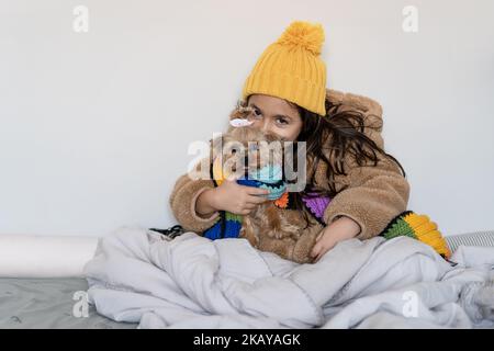 Enfant vêtu d'un manteau en fausse fourrure beige chaud et d'un bonnet et d'une écharpe tricotés assis avec un petit chien drôle dans un lit à la maison en hiver froid. Belle petite fille se réchauffe dans une couverture et regarde dans l'appareil photo. Banque D'Images