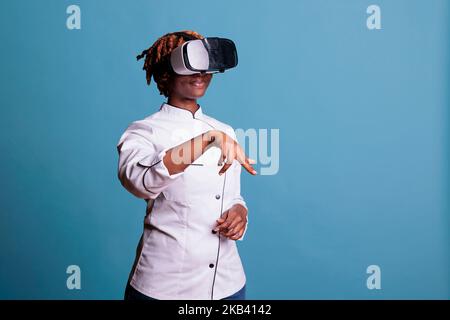 Chef féminin professionnel utilisant des lunettes de réalité virtuelle en studio. Cuisinier afro-américain portant l'uniforme excitamment dans une simulation de créations culinaires sur fond bleu. Banque D'Images