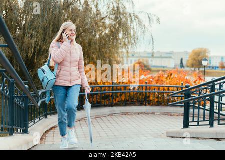 Une fille blonde parle au téléphone et se promène dans la ville sur fond de feuilles d'automne et rit par temps ensoleillé. Banque D'Images