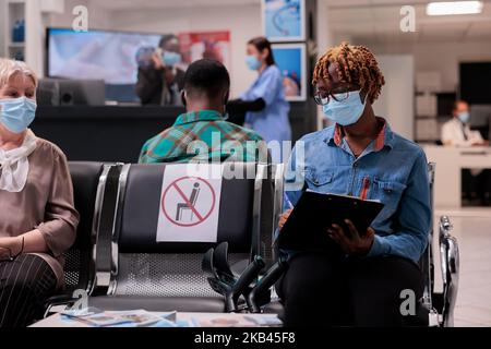 Patient afro-américain avec problème de mobilité remplissant le formulaire médical pour subir des tests. La chambre d'hôpital est bondée de personnes diverses portant des masques pour se protéger des covid. Banque D'Images