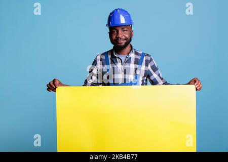 Optimiste constructeur en casque de sécurité tenant une grande bannière jaune vide pour l'industrie de la construction concept sur fond bleu. Constructeur avec combinaison présentant un panneau avec espace publicitaire. Banque D'Images