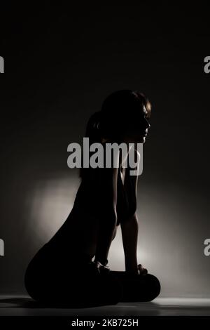 Posez la fille au repos et à l'étirement après avoir pratiqué des poses de yoga. Demi-silhouette avec éclairage latéral sur fond sombre. Banque D'Images