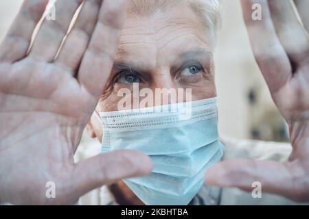Mains, covid et quarantaine avec un homme âgé dans un masque debout derrière le verre dans une maison pendant le verrouillage. Fenêtre, triste et isolé avec un homme mature Banque D'Images