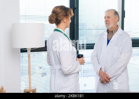 Portrait d'une femme médecin et d'un médecin chirurgien adulte principal ayant une conversation, buvant du café pendant la pause, debout sur le fond de la fenêtre. Banque D'Images