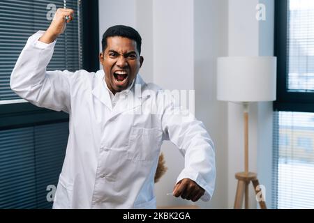 Un homme afro-américain fou en uniforme blanc criant furieux, criant avec colère tenant dans la seringue à main, regardant la caméra. Banque D'Images
