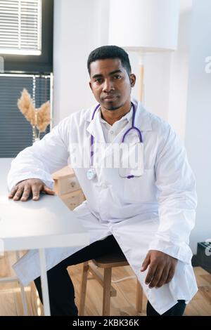 Portrait vertical d'un médecin afro-américain sérieux portant un manteau uniforme blanc avec stéthoscope assis à une table dans un cabinet médical. Banque D'Images