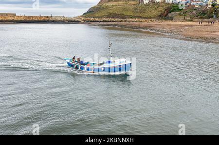 Un petit bateau de pêche entre dans un port. Une plage est d'un côté et une jetée est au loin. Banque D'Images