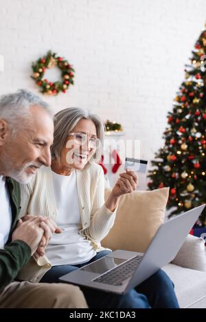 heureux homme mature près de la femme avec carte de crédit regardant l'ordinateur portable tout en ayant des achats en ligne pendant noël Banque D'Images