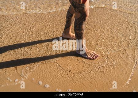 La vue en grand angle des jambes tatouées d'un homme debout sur le sable d'une plage Banque D'Images