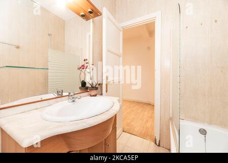 Salle de bains avec meuble en merisier avec miroir intégré, comptoir en marbre crème, sol en grès et cabine de douche avec cloison en verre Banque D'Images