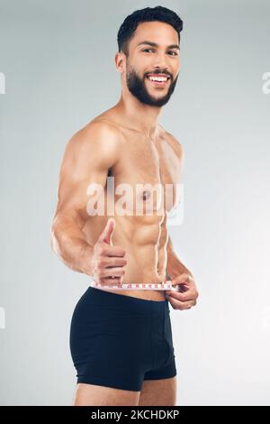 Corps, taille et pouces vers le haut avec un modèle d'homme en studio sur un fond gris pour promouvoir la perte de poids ou l'exercice. Portrait, forme physique et régime avec un jeune Banque D'Images