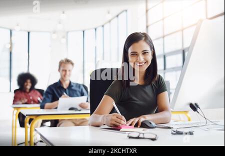 Écrivez votre propre script à la réussite. Portrait d'un designer heureux assis à son bureau pendant que ses collègues travaillent en arrière-plan. Banque D'Images