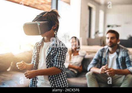 Donner au mot supervision une nouvelle signification. Un petit garçon utilisant un casque de réalité virtuelle à la maison avec ses parents en arrière-plan. Banque D'Images