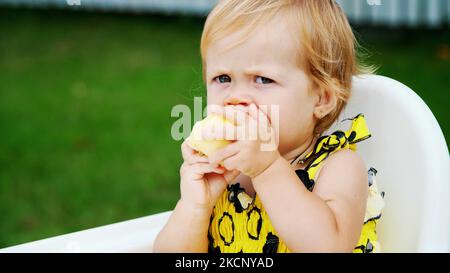 été, dans le jardin. portrait, une fille blonde d'un an mangeant une pomme avec de l'apetite. sur la table. Photo de haute qualité Banque D'Images