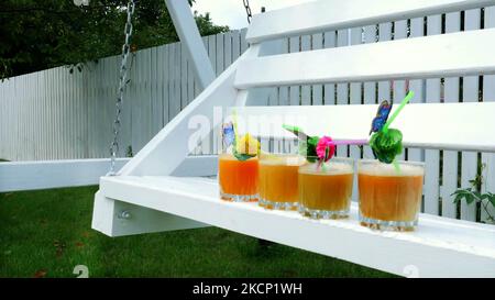 gros plan, sur une balançoire en bois blanc, dans le jardin, en été, 4 verres avec jus d'orange fraîchement pressé et de pêche sont debout, décorés de pailles pour cocktails. Photo de haute qualité Banque D'Images