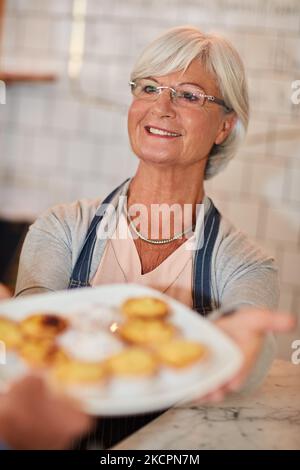 Prenez-les à la table deux, s'il vous plaît. Un propriétaire d'affaires senior heureux qui donne une assiette de pâtisseries à un serveur dans son café. Banque D'Images