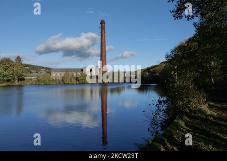 Grande cheminée d'usine de brique rouge avec réflexion dans le petit barrage de moulin lac avec arbres bâtiments ciel bleu et nuages blancs Banque D'Images