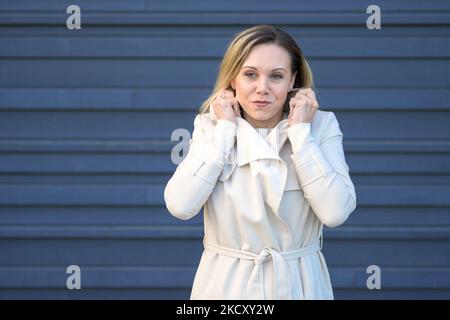 Belle femme blonde d'âge moyen tenant le col de son manteau avec ses mains car il fait très froid dehors devant un mur bleu Banque D'Images