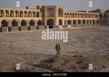 Une iranienne marche le long d'un lit de rivière Zayandeh séché dans la ville d'Ispahan, à 450 km (281 miles) au sud de Téhéran sur 14 décembre 2021 au coucher du soleil, comme une vue sur le pont historique de Khaju est vue sur la gauche. Zayandeh Rud est l'une des principales attractions touristiques d'Ispahan, qui a complètement séché, aussi les ponts historiques sur la rivière peuvent être endommagés en raison de l'affaissement du lit de la rivière Zayandeh Rud si la sécheresse continue. (Photo de Morteza Nikoubazl/NurPhoto) Banque D'Images