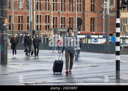 La vie quotidienne avec des gens et un passager avec des bagages regardant sa montre comme vu avec la gare centrale d'Amsterdam en arrière-plan. Les habitants et quelques touristes dans les rues calmes d'Amsterdam pendant le confinement dans la capitale néerlandaise avec des magasins et des magasins paraissant avec fermé avec le roller métal volet vers le bas, cafés, bars et restaurants également fermés avec des tables et des chaises des terrasses fermées. Les pays-Bas ont été la première nation européenne à déclarer un verrouillage complet pour lutter contre la nouvelle variante d'Omicron qui s'intensifie. Après un ordre soudain du gouvernement avant Noël, le pays a fermé Banque D'Images