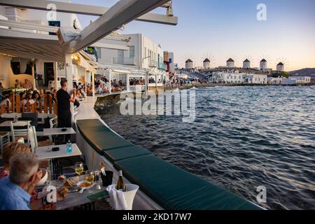 Les moulins à vent emblématiques de l'île de Mykonos vus de la petite Venise avec des touristes dans les bars, cafés et restaurants. Mykonos est une île en Grèce, dans la mer Égée. Il y a 16 moulins à vent sur l'île, dont 5 au-dessus de Chora, la ville principale de l'île. Les moulins à vent ont été construits au 16th siècle à partir des Vénitiens mais leurs constructions se sont poursuivies jusqu'au 20th siècle. Coucher de soleil avec les gens aux moulins à vent de Mykonos. Les touristes apprécient un beau coucher de soleil pendant l'heure magique à l'île de Mykonos en Grèce. L'île grecque de Myconos est une destination de voyage populaire et glamour pour la Méditerranée Banque D'Images