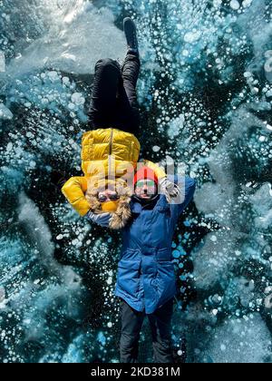 Heureux couple couché sur le lac Baikal d'hiver gelé portant des vêtements d'hiver. Glace transparente. Voyage en hiver, loisirs actifs, sports, vacances. Banque D'Images