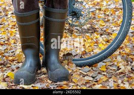 Avec des pieds secs sur le vélo jusqu'à l'automne. Le cycliste est debout dans des bottes en caoutchouc dans un feuillage coloré devant un pneu de vélo. Banque D'Images