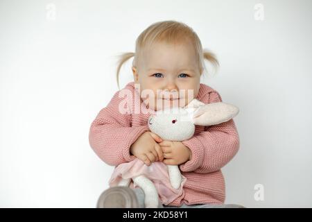 Belle petite fille jouer avec animal bourré, isolé sur blanc. Un tout-petit joyeux se fait un plaisir d'embrasser un lapin en peluche. Enfant blond aux cheveux vêque de vêtements roses Banque D'Images