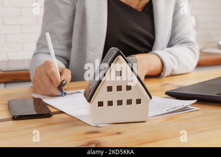 Une femme d'affaires signe des documents sur l'arrière-plan d'un modèle de maison contre l'arrière-plan de meubles de cuisine. Le concept d'acheter une maison, Mo Banque D'Images