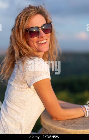 Bonne jeune femme de race blanche positive dans des lunettes de soleil riant au coucher du soleil à l'extérieur Banque D'Images