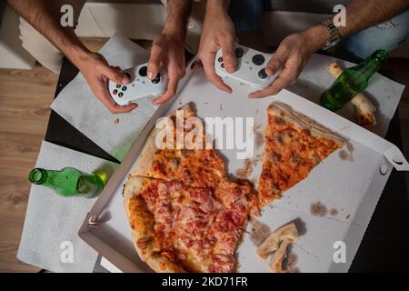 Deux amis tenant leurs manettes de jeu jouant à des jeux vidéo et mangeant de la pizza. Banque D'Images