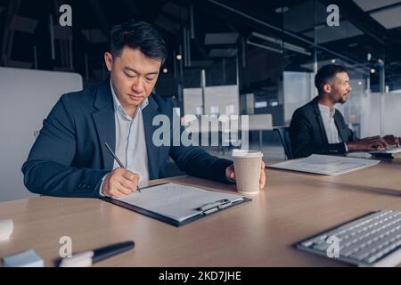 Homme d'affaires asiatique concentré qui boit du café tout en travaillant avec des documents dans un bureau moderne Banque D'Images