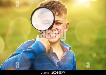 J'espionne avec mon petit œil... Portrait d'un petit garçon mignon regardant à travers une loupe tout en se tenant dehors. Banque D'Images