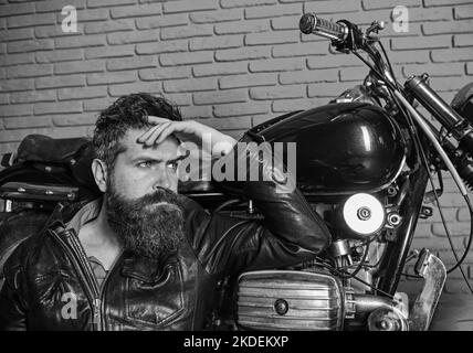 Concept de style de vie des motards. Homme avec la barbe, motard dans la veste en cuir près de moto dans le garage, arrière-plan mural en brique. Taille basse, motard brutal sur le pensif Banque D'Images