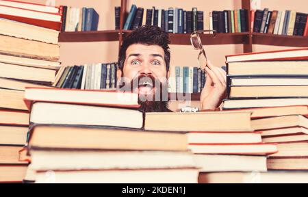 Homme sur le visage heureux entre des piles de livres dans la bibliothèque, des étagères sur le fond. Concept de recherche scientifique. Le professeur ou l'élève porte une barbe Banque D'Images