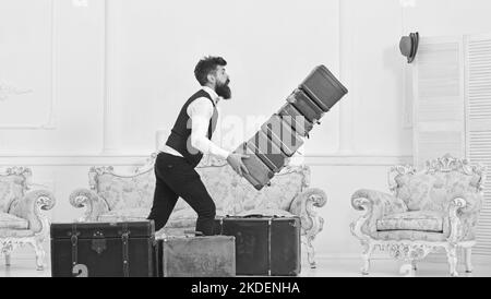 Concept d'assurance bagages. Homme avec barbe et moustache en costume classique livre des bagages, luxe intérieur blanc arrière-plan. Porter, maître d'hôtel accidentellement Banque D'Images